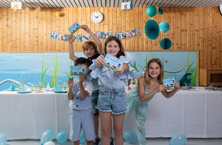 4 Kinder (2 Jungen und 2 Mädchen) stehen nebeneinander und halten Einladungskarten für einen Kindergeburtstag in Linz AG Bädern in die Kamera. Dahinter befindet sich ein Geburtstagstisch und eine Happy Birthday Girlande.